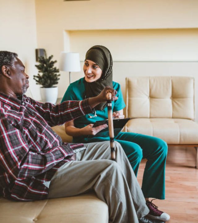 A Muslim female doctor is visiting her older patient of Afro-American ethnicity. An Arabian female healthcare worker is helping her older male patient in the living room at home.